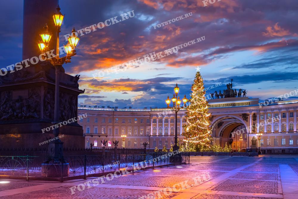 На фотографии изображена вечерняя Дворцовая площадь в Санкт-Петербурге зимой. Праздничная елка украшает центр площади, а на фоне виден Главный штаб с подсветкой. Уличные фонари и закатное небо создают атмосферу волшебства и праздника. Этот вид передает зимнее настроение и красоту новогоднего Петербурга.